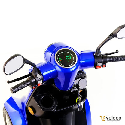 Na sliki je električni skiro znamke Veleco, model Gravis. Vidna je sprednja stran skiroja, kjer so prikazani krmili, merilnik hitrosti in nivoja baterije ter ogledali. Barva skiroja je modra, kar mu daje privlačen videz. Na krmilu so tudi različni gumbi za upravljanje.