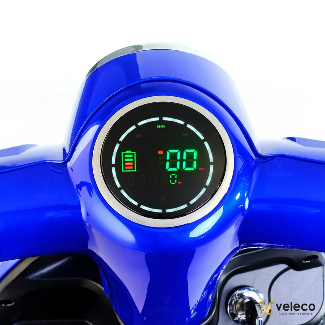 Izdelek je električni skiro, znamke Veleco, model Gravis. Na sliki je prikazan del krmila z digitalnim zaslonom, ki prikazuje informacije o bateriji in hitrosti. Barva skiroja je modra, zaslon pa ima jasno berljive podatke, kar omogoča enostavno spremljanje delovanja vozila.