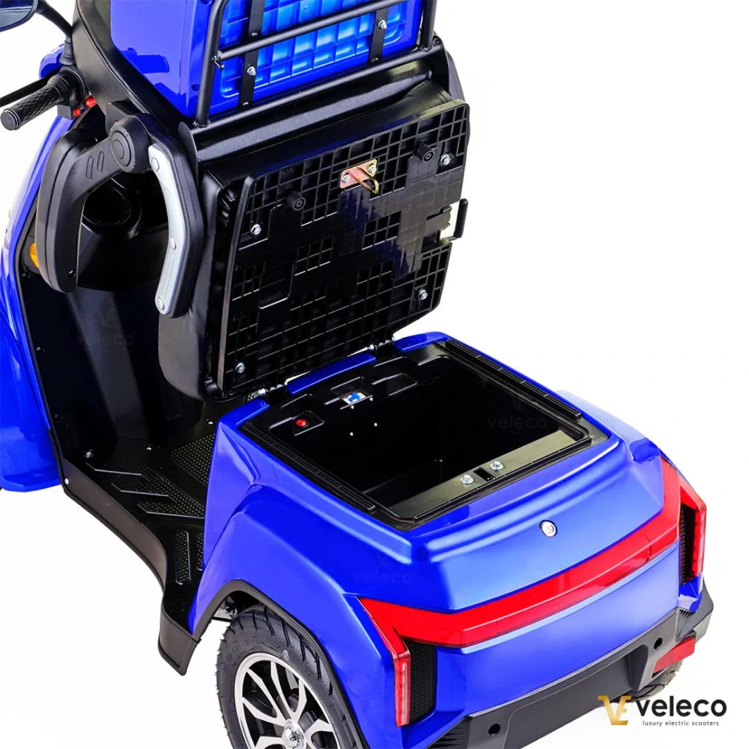 Izdelek je električni skuter, model Veleco Gravis. Na sliki je prikazana zadnja stran skuterja v modri barvi, ki ima odprt prostor za shranjevanje. Vidna je tudi zasnova koles in detajli na karoseriji, kar nakazuje na funkcionalnost in udobje pri vožnji.