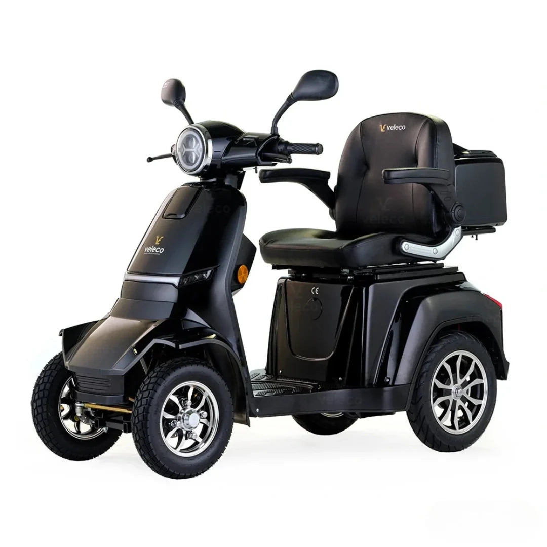 Izdelek na sliki je električni skuter, natančneje model Veleco Gravis. Na sliki je prikazan skuter v črni barvi, opremljen s širokim sedežem in prostornim prostorom za shranjevanje. Ima tri kolesa, kar zagotavlja stabilnost, ter svetlobno napravo na sprednjem delu. Skuter je zasnovan za udobno vožnjo in enostavno upravljanje.