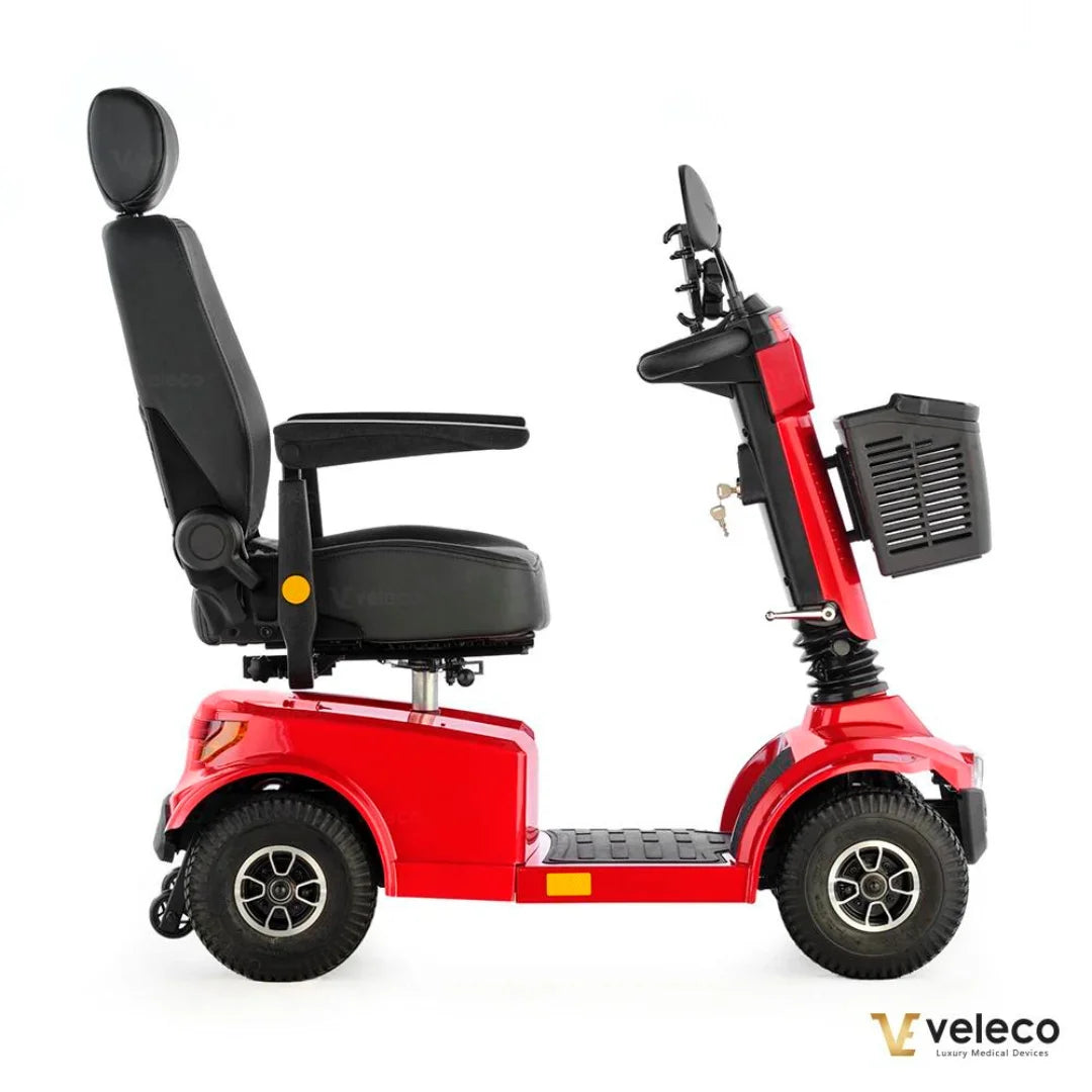 Izdelek je električni skuter, model Veleco Jumpy. Na sliki je prikazan stranski pogled na skuter, ki ima udobno sedežno mesto z naslonjalom in naslonoma za roke. Skuter je v svetlo rdeči barvi in ima široke pnevmatike, kar omogoča stabilno vožnjo. Na zadnji strani je vidna tudi košara za shranjevanje.