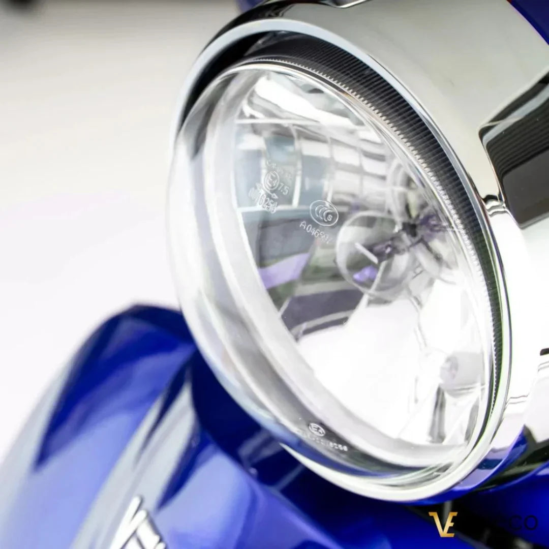 Izdelek je električni mobilni voziček, model Veleco ZT15. Na sliki je prikazana sprednja luč vozička, ki ima kromiran rob in prozorno steklo. Barva vozička je modra, kar je vidno na ozadju. Luč je zasnovana za zagotavljanje vidnosti in varnosti med vožnjo.