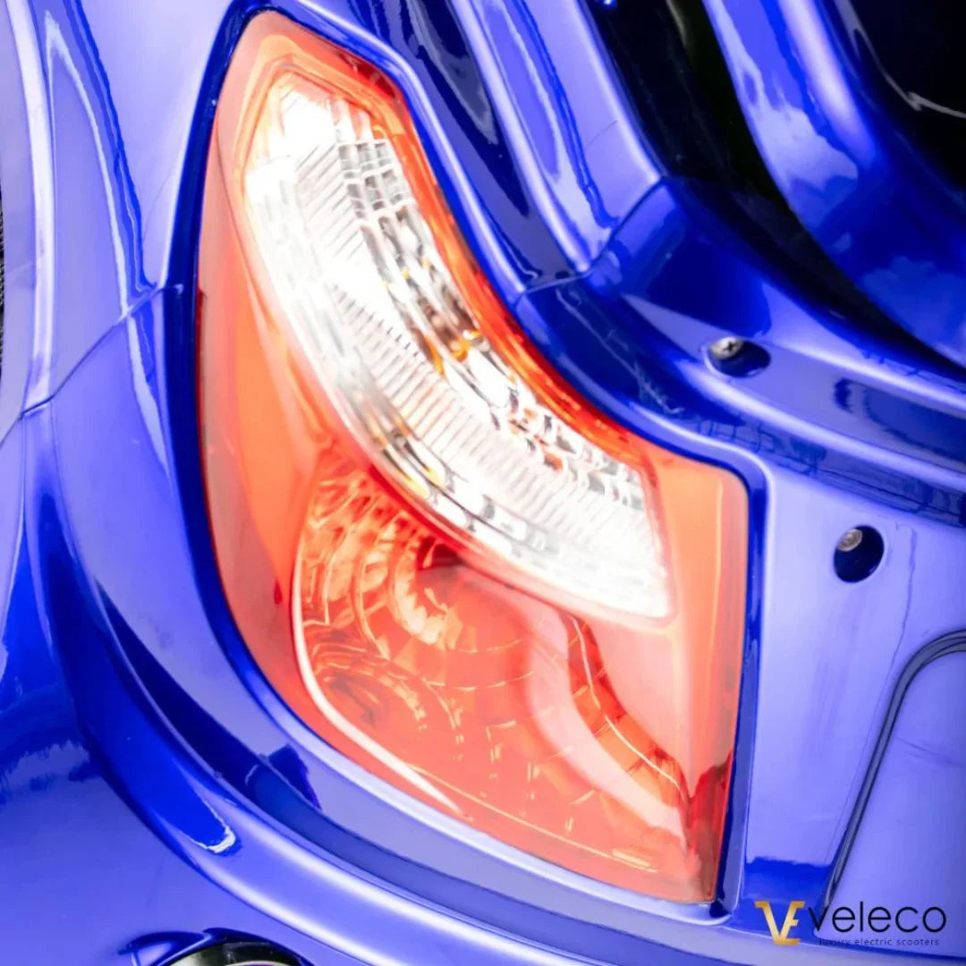 Izdelek je električni mobilni voziček, model Veleco ZT15. Na sliki je vidna zadnja luč vozička, ki je v modri barvi. Luč ima kombinacijo rdeče in prozorne plastike, kar je značilno za signalizacijo pri električnih vozilih. Opremljen je s sodobnim dizajnom, kar daje vozičku privlačen videz.