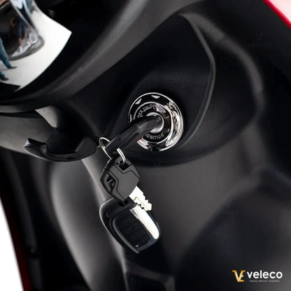 Izdelek je električni mobilni voziček, model Veleco ZT63. Na sliki je prikazan ključavnica za vžig, ki je del nadzorne plošče vozička. Ključ je vključen in vidna je tudi bližnja ročica za udobno držanje. Barve in materiali so sodobni, kar kaže na kakovost izdelka.