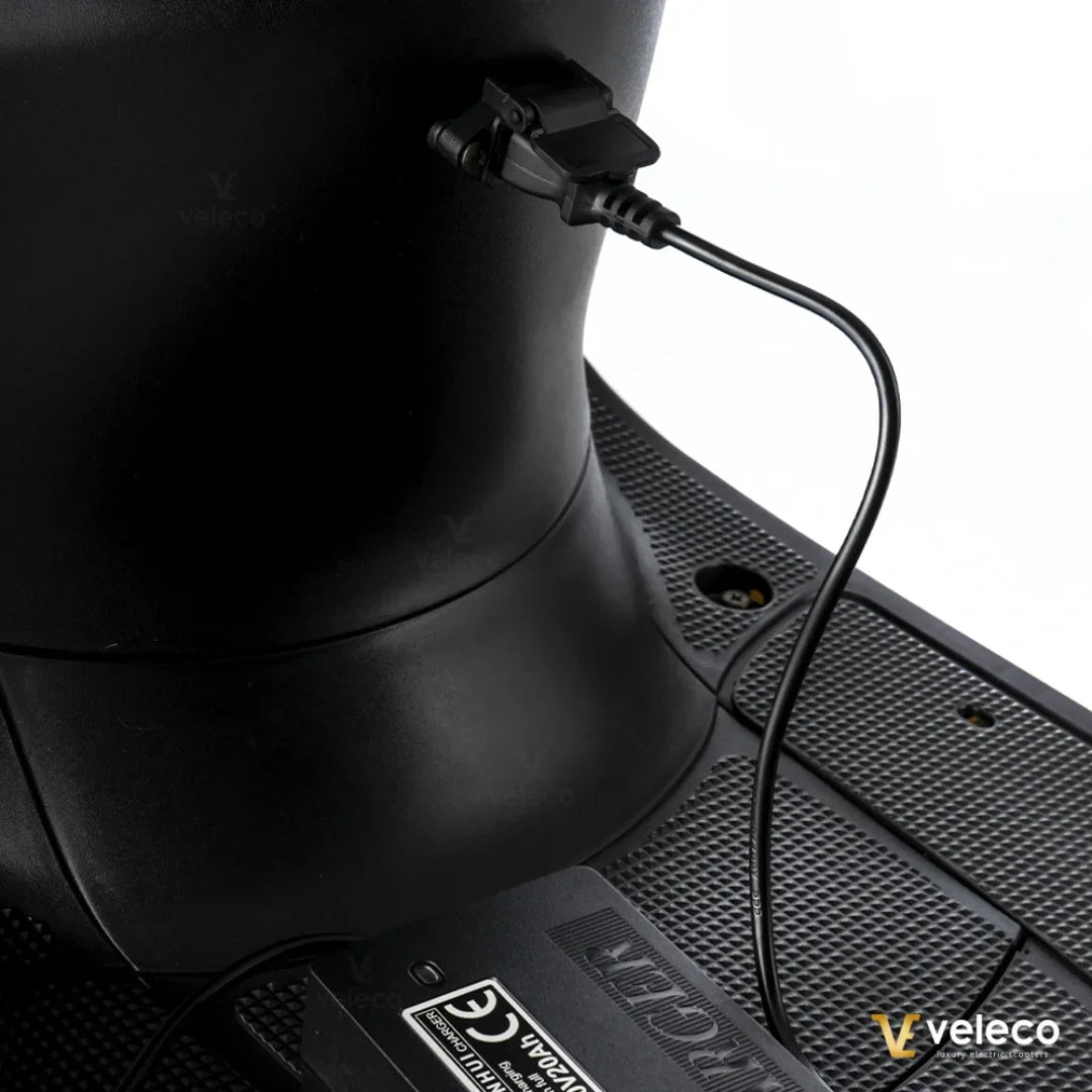 Izdelek je električni mobilni voziček, model Veleco ZT63. Na sliki je prikazan del vozička, kjer je vidna povezava za polnjenje, ki je povezana z električnim sistemom. Površina vozička je narejena iz materiala, ki nudi dober oprijem, kar je pomembno za varno uporabo.