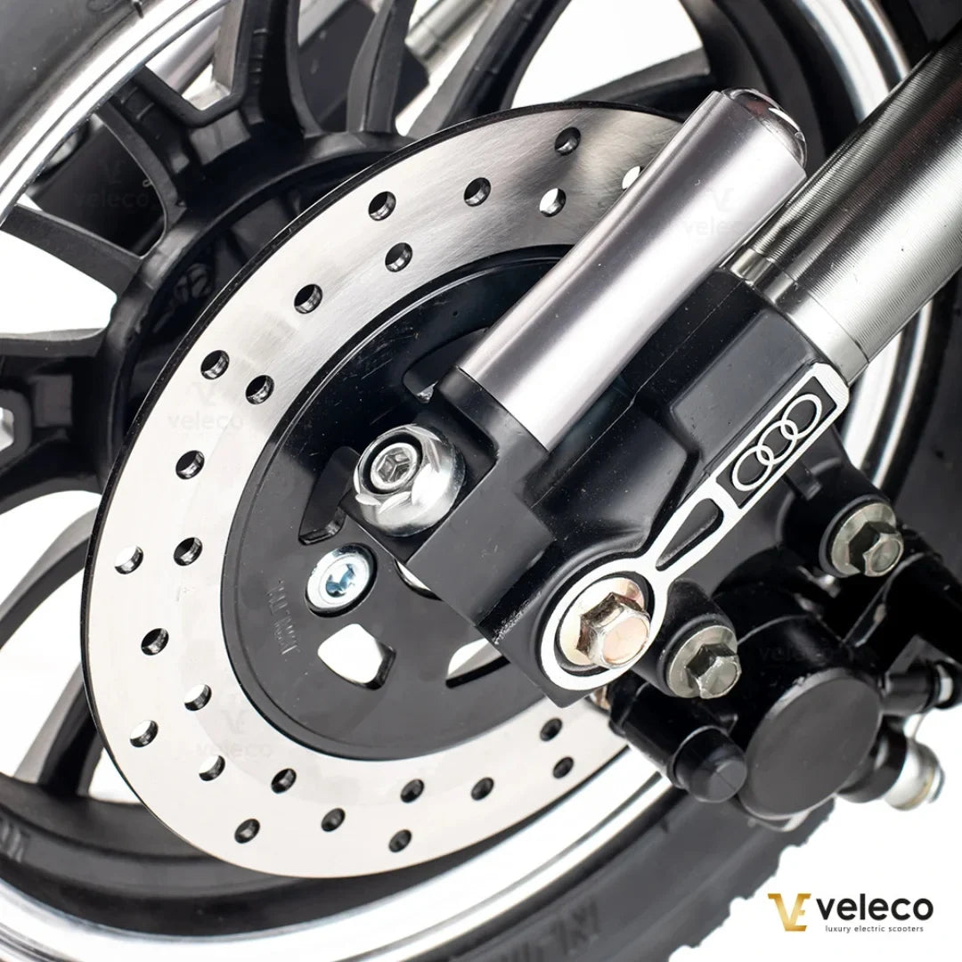 Izdelek je električni mobilni voziček, model ZT63, znamke Veleco. Na sliki je prikazan del kolesa, ki vključuje disk zavoro in del mehanizma za zaviranje. Vidna je tudi aluminijasta cev in del kolesa, kar nakazuje na robustno in kakovostno zasnovo.