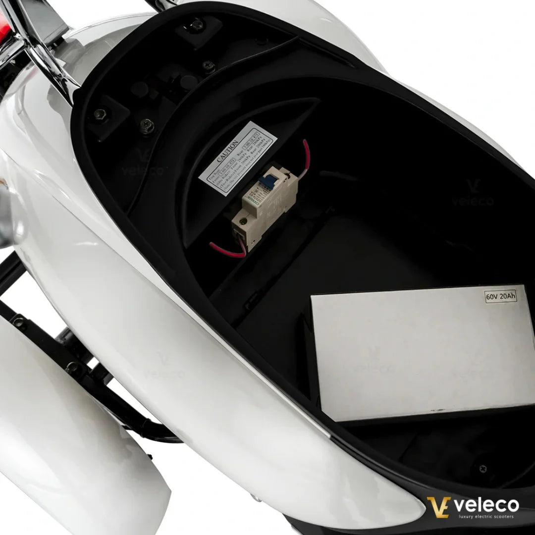 Izdelek je električni mobilni voziček, model Veleco ZT63. Na sliki je prikazan prostor za shranjevanje, kjer se nahaja baterija in električni varovalke. Barva vozička je bela, kar daje sodoben in eleganten videz.
