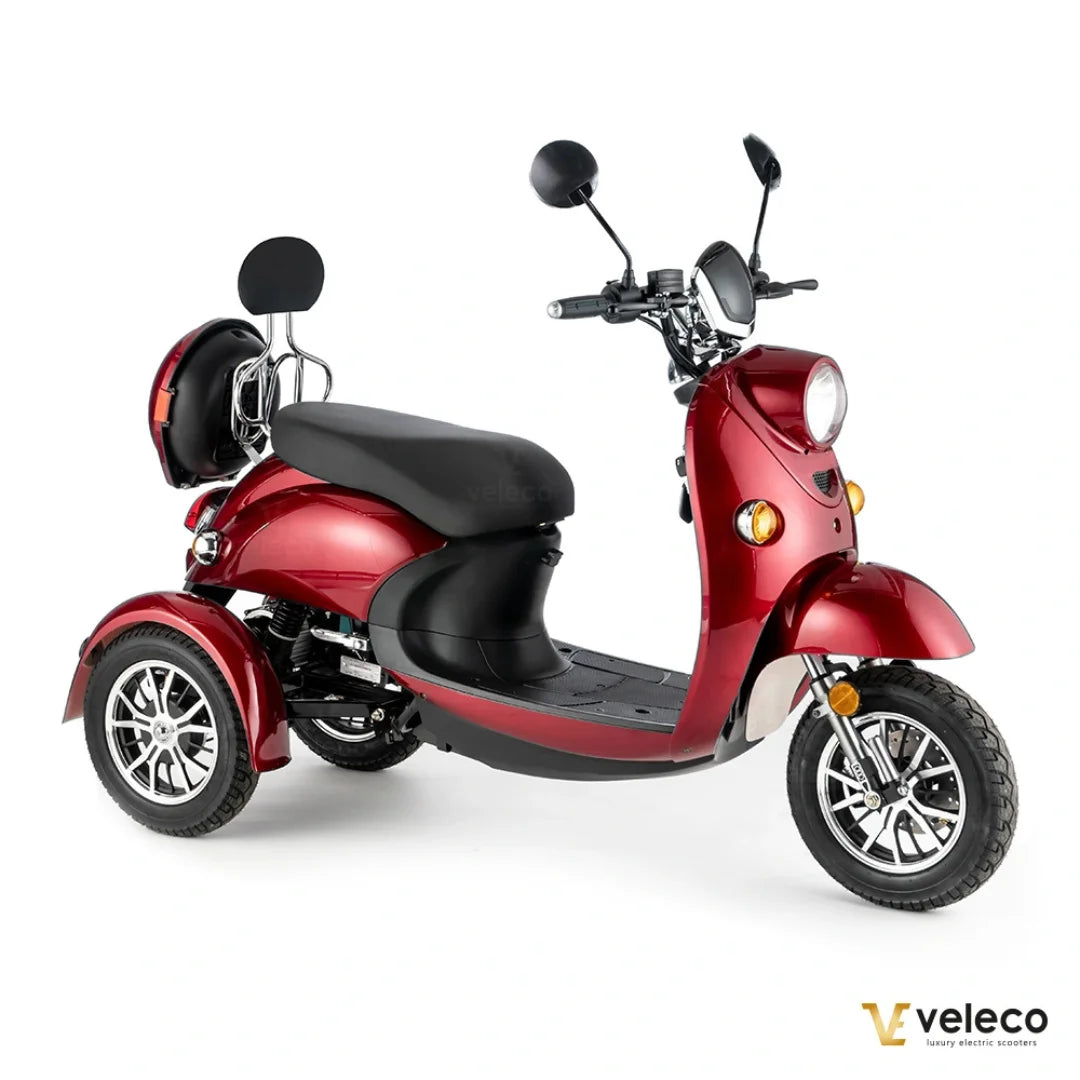 Izdelek je električni mobilni voziček, model Veleco ZT63. Na sliki je prikazan voziček v rdeči barvi, ki ima tri kolesa, kar omogoča stabilno vožnjo. Ima široko sedežno površino, sprednje luči in ogledala, kar ga naredi primernega za mestno vožnjo. Na zadnjem delu je nameščen prtljažnik, kar povečuje njegovo funkcionalnost.