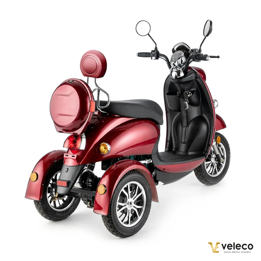 Izdelek na sliki je električni mobilni voziček, model Veleco ZT63. Ima tri kolesa, kar zagotavlja stabilnost, in je zasnovan za udobno vožnjo. Na sliki je vidna elegantna rdeča barva, široko sedežno mesto ter krmilo s ogledali. Voziček je opremljen z zadnjim prtljažnikom in lučmi, kar povečuje njegovo funkcionalnost in varnost.