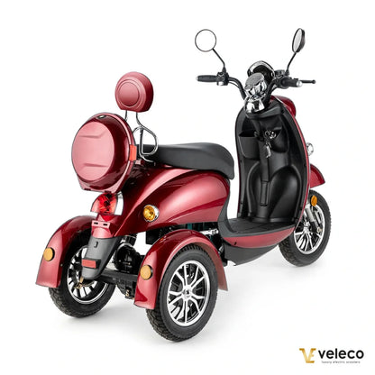 Izdelek na sliki je električni mobilni voziček, model Veleco ZT63. Ima tri kolesa, kar zagotavlja stabilnost, in je zasnovan za udobno vožnjo. Na sliki je vidna elegantna rdeča barva, široko sedežno mesto ter krmilo s ogledali. Voziček je opremljen z zadnjim prtljažnikom in lučmi, kar povečuje njegovo funkcionalnost in varnost.