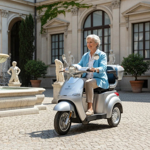 Izdelek na sliki je električni skuter, model Veleco Cristal. Na sliki vidimo starejšo žensko, ki se vozi s skuterjem v slikovitem okolju, obdanem s klasično arhitekturo in skulpturami. Skuter je srebrne barve in ima udobno sedežno površino ter prostoren zadnji predal.