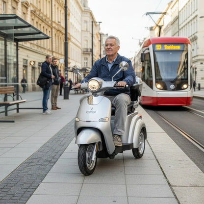 Izdelek na sliki je električni skuter, model Veleco Cristal. Na sliki je videti moškega, ki vozi skuter po urbanem okolju, ob njem pa se nahaja tramvaj in pešci. Skuter ima sodoben dizajn in je opremljen s svetlobnimi elementi, kar daje občutek udobja in mobilnosti v mestnem prometu.