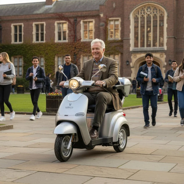 Na sliki je električni skuter znamke Veleco, model Cristal. Izdelek je srebrne barve in ima retro obliko. Na skuterju sedi starejši moški, ki se nasmejan vozi po dvorišču univerze, obkrožen z mladimi ljudmi, ki se sprehajajo. V ozadju so vidne stavbe z značilno arhitekturo, kar daje sliki prijeten in živahen ambient.