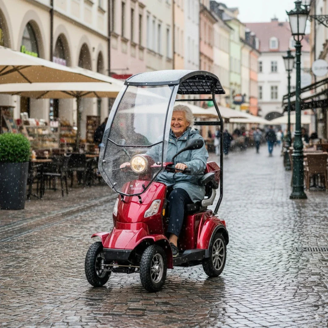 Na sliki je električni mobilni skuter, model Veleco Faster. Izdelek je v rdeči barvi in ima zaščitni pokrov, ki ščiti voznika pred dežjem. Na skuterju sedi starejša ženska, ki se nasmejana vozi po tlakovanem mestu, obdanem s trgovinami in kavarnami. V ozadju je vidna urbana pokrajina, kar daje občutek živahnosti in aktivnosti.