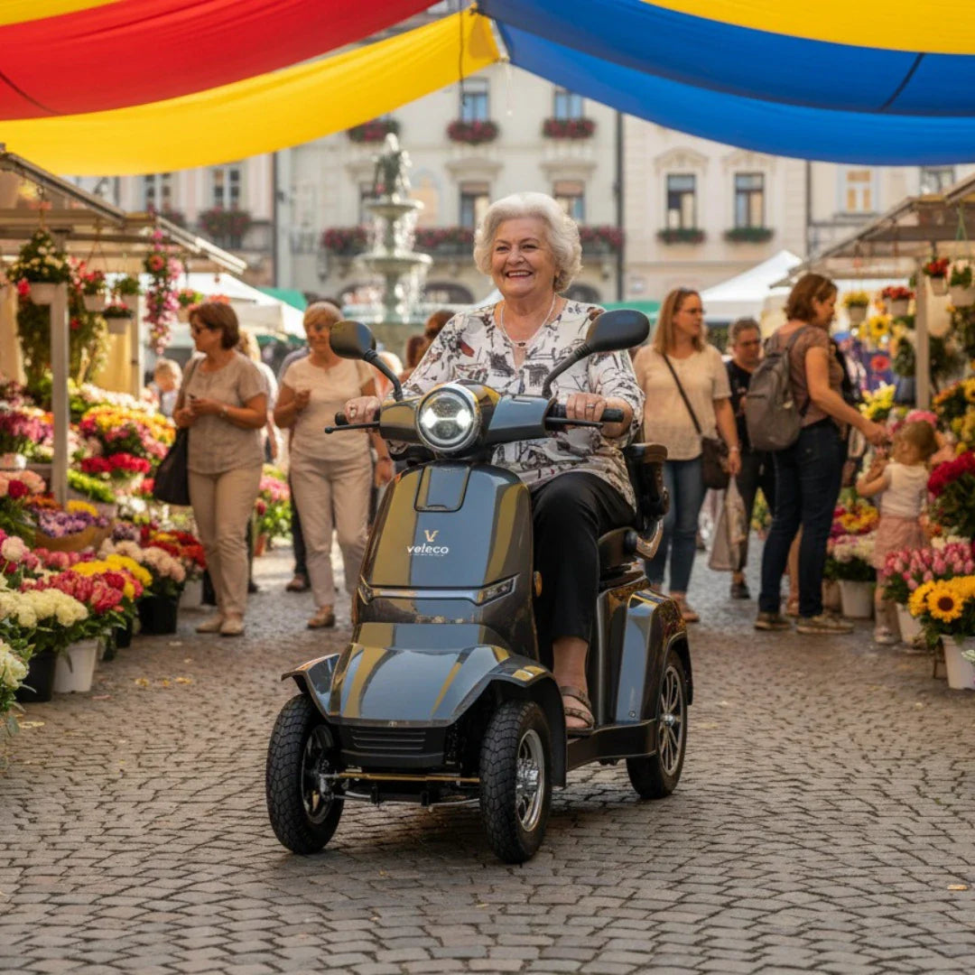 Izdelek na sliki je električni skuter, model Veleco Gravis. Na sliki vidimo starejšo žensko, ki se smehlja med vožnjo po tržnici, obdani s pisanimi cvetlicami in barvitimi strešnimi pokrivali. Okolica je živahna, z več ljudmi, ki uživajo v nakupovanju. Skuter ima tri kolesa, kar zagotavlja stabilnost in udobje pri vožnji.