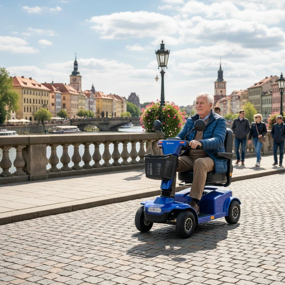 Izdelek je električni skuter, model Veleco Jumpy. Na sliki je videti starejša oseba, ki vozi skuter po urejeni pešpoti ob reki. Ozadje prikazuje slikovite stavbe in zelenje, kar ustvarja prijetno urbano okolje. Skuter je modre barve in ima udoben sedež ter košarico spredaj.
