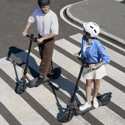 Na sliki je prikazan električni skiro znamke Xiaomi, model Elite. Dva uporabnika, moški in ženska, se peljeta po prehodu za pešce. Oba nosita čelade za zaščito, kar nakazuje na varno vožnjo. Ozadje vključuje asfaltirano cesto in peščen prehod, kar ustvarja urbano okolje.