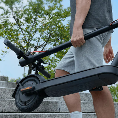 Na sliki je električni skiro znamke Xiaomi, model Elite. Oseba ga drži v roki, medtem ko hodi po stopnicah. Skuter ima črno zasnovo, široke gume in ergonomsko oblikovan ročaj, kar nakazuje na udobno vožnjo. V ozadju so vidne drevesne krošnje in delno oblačno nebo.