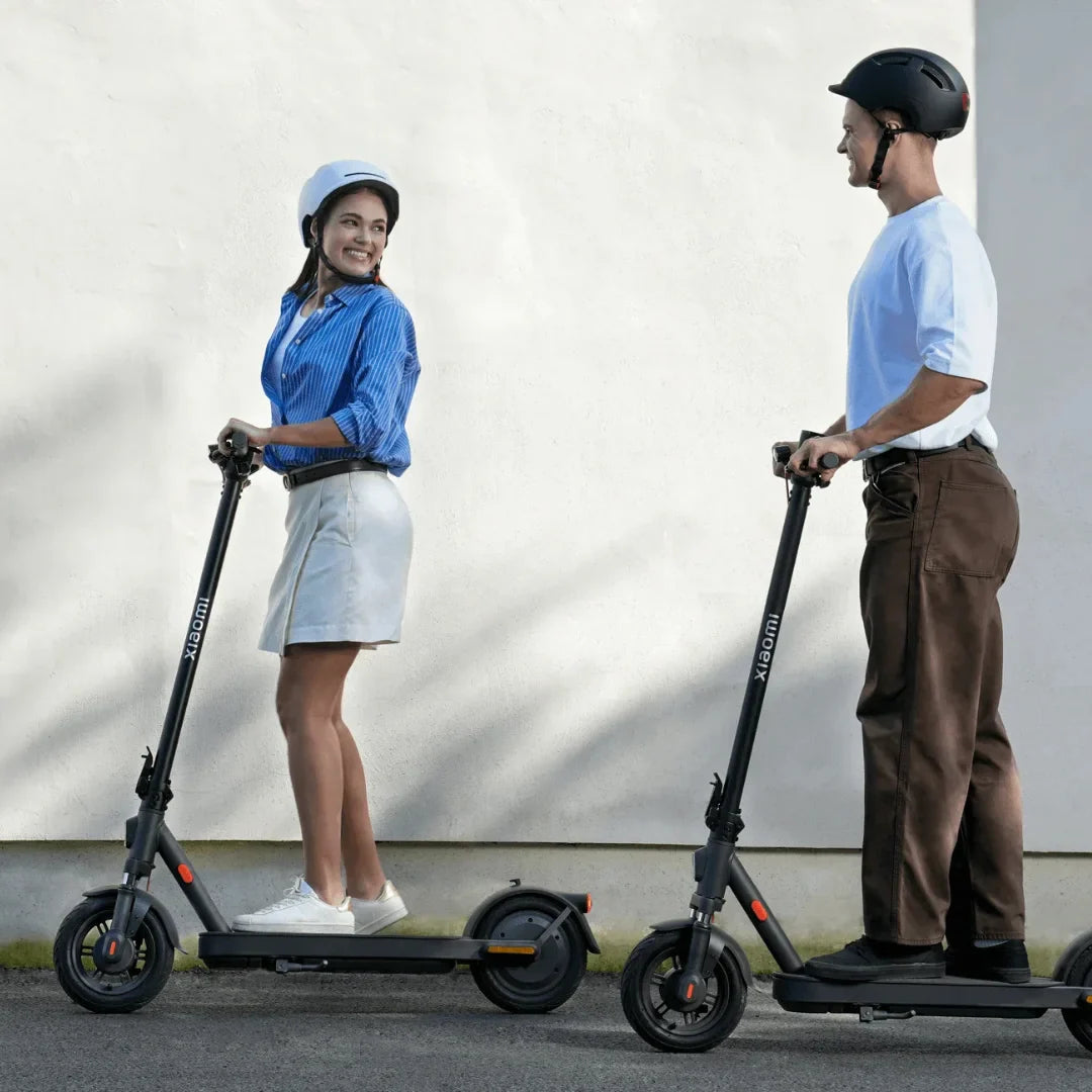 Na sliki je prikazan električni skiro znamke Xiaomi, model Elite. Dva uporabnika, moški in ženska, se vozita na skirojih po urbanem okolju. Oba nosita čelade za zaščito, ženska je oblečena v modro srajco in belo krilo, moški pa v belo majico in rjave hlače. Ozadje je preprosto, s svetlo steno, kar poudarja skiroja.