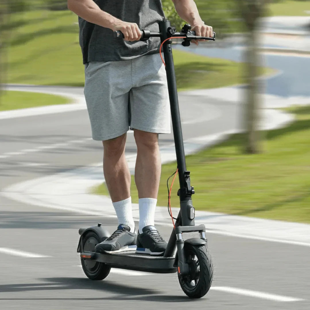 Na sliki je električni skiro znamke Xiaomi, model Elite. Oseba na skiroju vozi po cesti, obkrožena z zelenjem. Ima na sebi športno obleko, vključno s kratkimi hlačami in športnimi copati. Skuter ima moderno zasnovo z velikimi kolesi in udobno platformo za stojo.