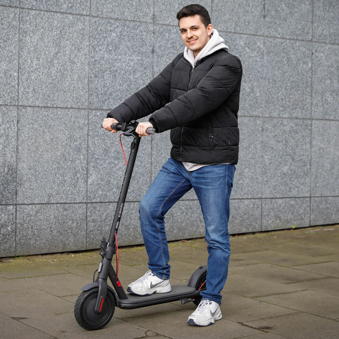 Oseba v črni jakni vozi električni skiro Xiaomi 4 Eu ob sivi steni.