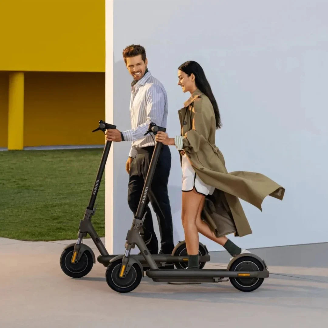 Na sliki je prikazan električni skiro, model Xiaomi 5 Max GL. Dva človeka, moški in ženska, se sprehajata in držita skiroje, ki sta v nevtralni sivi barvi. Obe osebi sta oblečeni v sproščena oblačila, okolica pa je moderna in svetla, kar ustvarja prijetno vzdušje.