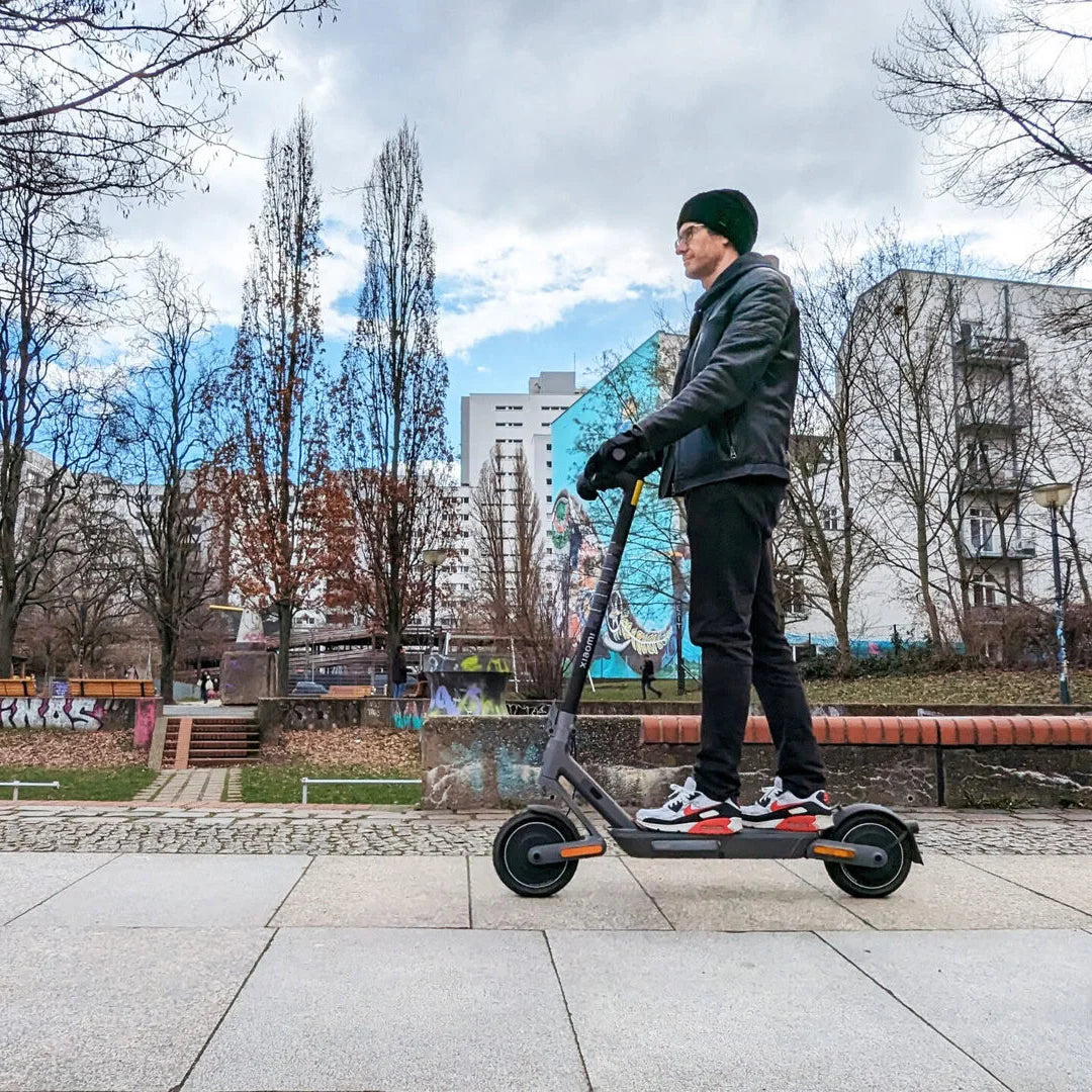 Oseba se vozi z električnim skirojem Xiaomi 4 Ultra po mestnem pločniku z drevesi in stavbami.