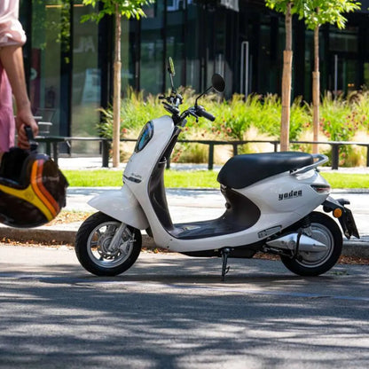 Bel električni skuter Yadea C Umi parkiran na mestni ulici z zelenjem v ozadju.