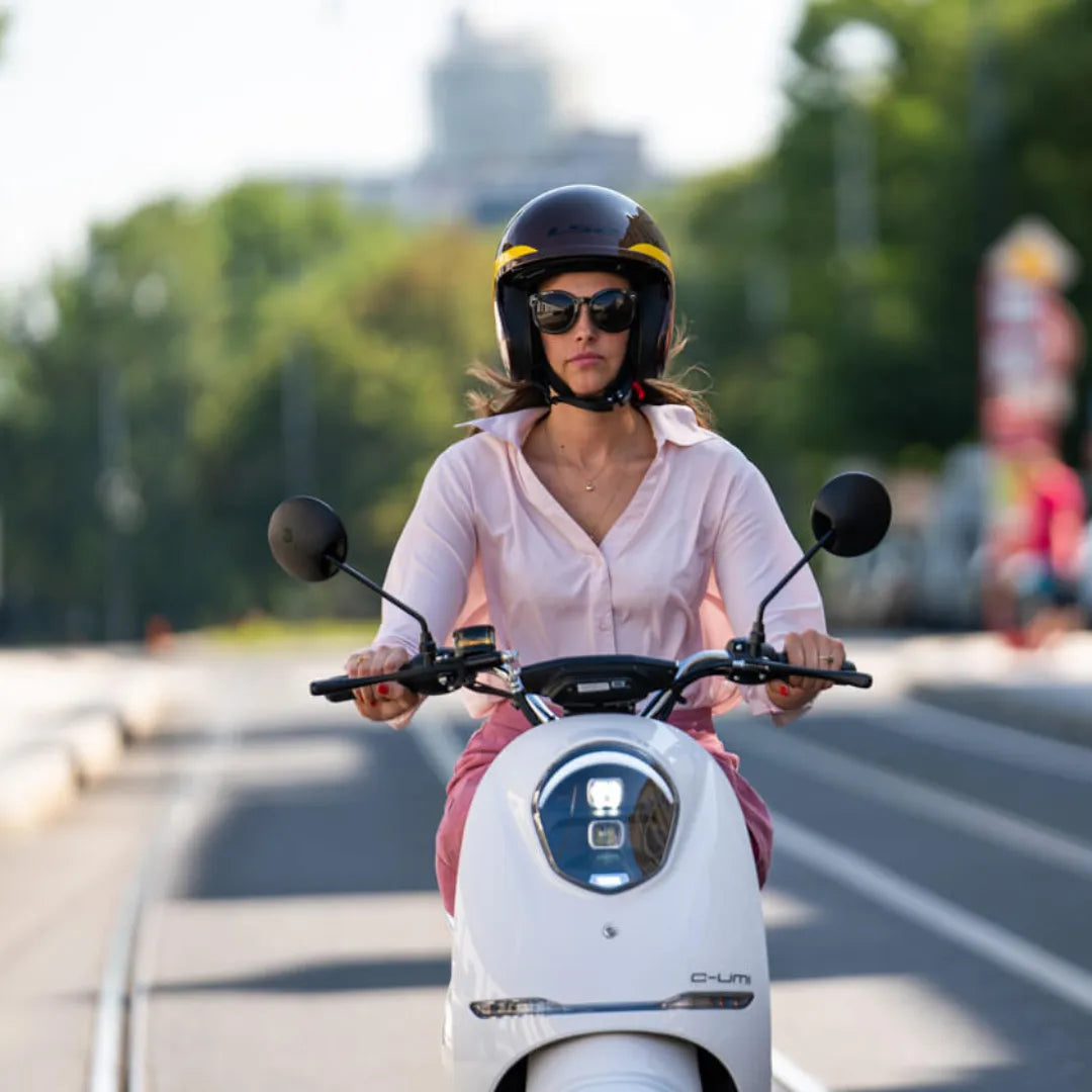 Ženska vozi električni skuter Yadea C Umi po mestni ulici, pri tem pa nosi čelado in sončna očala.