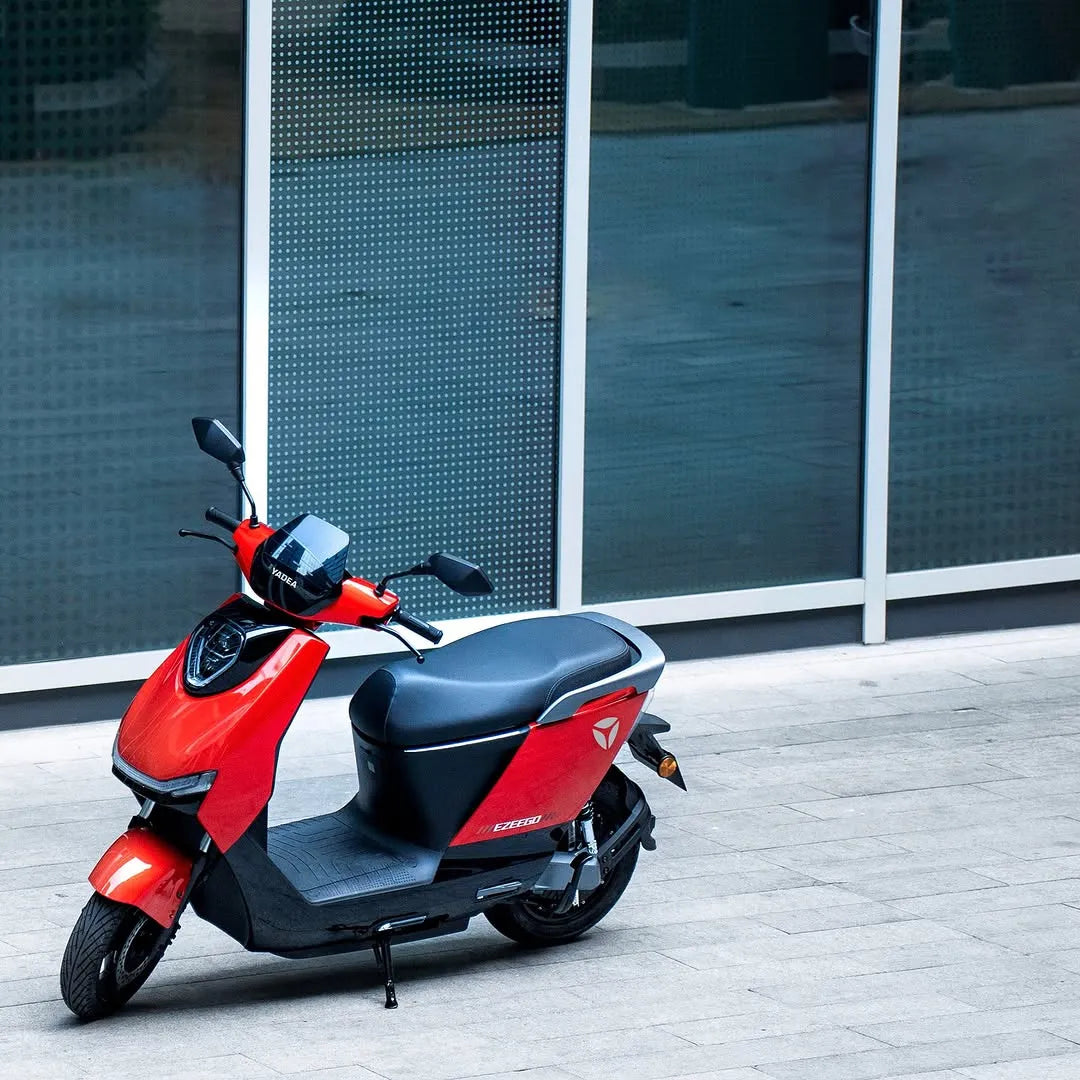 Rdeč električni skuter Yadea Ezeego parkiran blizu moderne steklene stavbe.