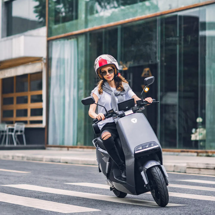 Ženska vozi električni skuter Yadea G5 po mestni ulici, nosi čelado in elegantno opravo.