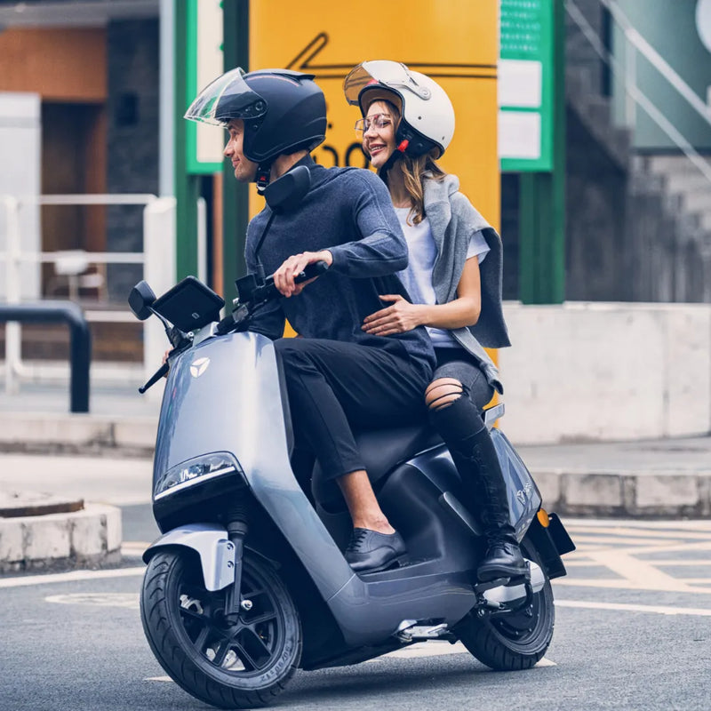 Par se vozi z električnim skuterjem Yadea G5 po mestni ulici, oba nosita čelado.
