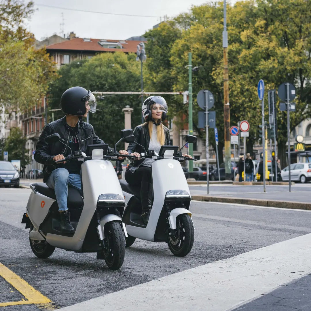 Dva voznika na električnem skuterju Yadea G5, ki se vozita po mestni ulici z drevesi v ozadju
