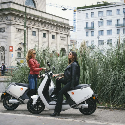 Dve ženski v elegantnih jaknah stojita ob električnem skuterju Yadea G5 v urbanem okolju.