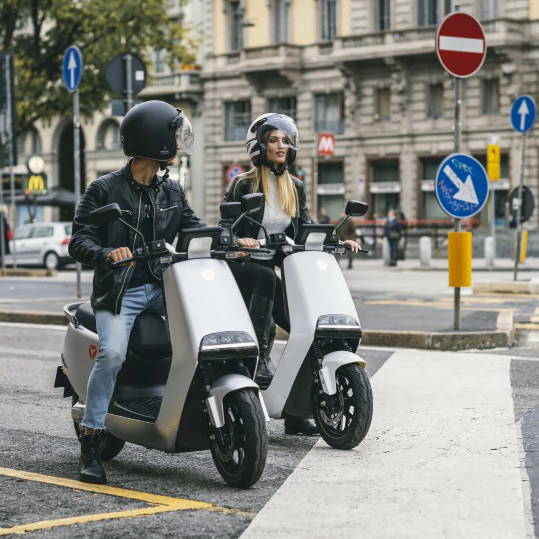 Dva voznika na električnem skuterju Yadea G5 na mestnem križišču s prometnimi znaki.