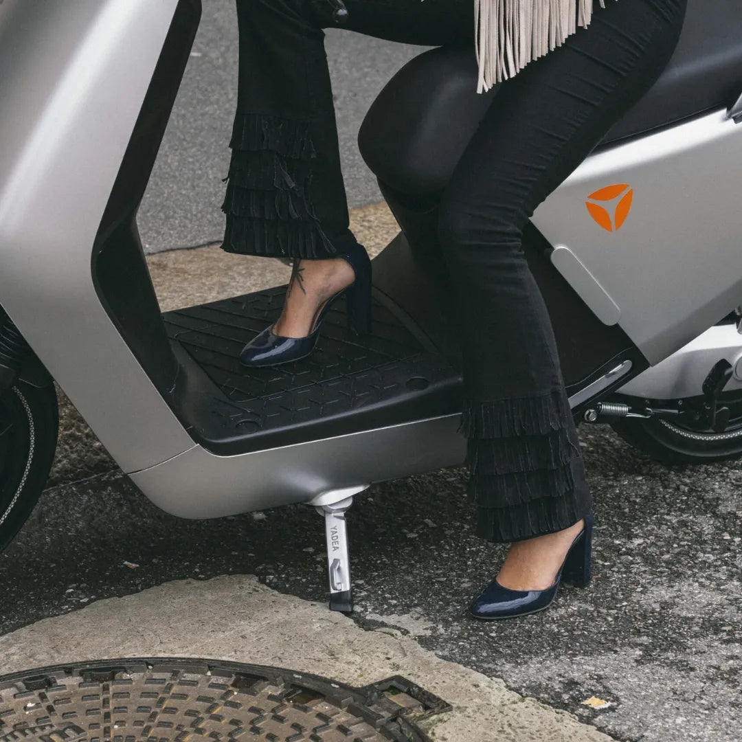 Oseba v elegantnih hlačah z resami in petah sedi na električnem skuterju Yadea G5.